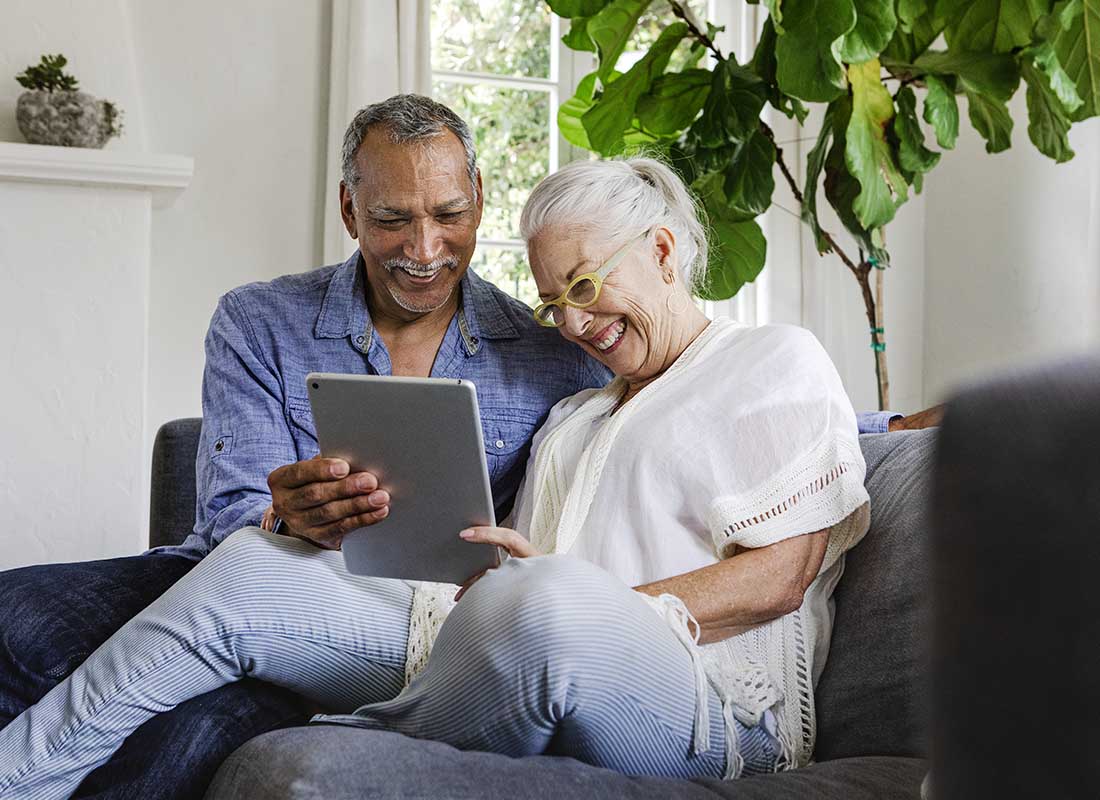 Medicare Supplement Insurance - Senior Couple Laughing Together and Using a Tablet While Sitting on a Couch in Their Home